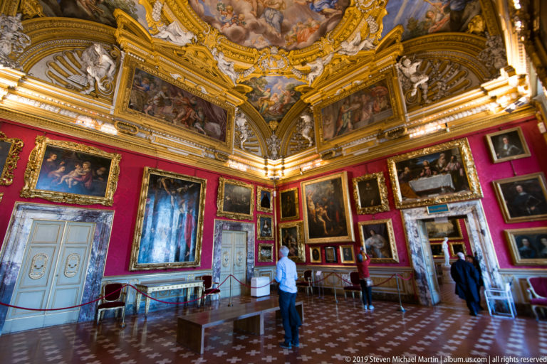 Visitors stand among densely hung paintings beneath gilded vaulted frescoes in a large gallery with patterned floor.