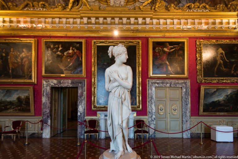 White marble female statue centered before a red gallery wall hung with large framed paintings and marble-trimmed doors.