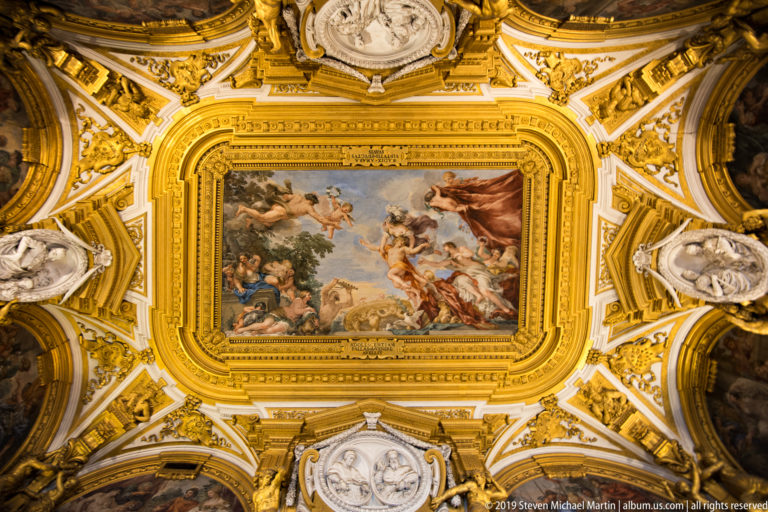 Upward view of a central mythological fresco surrounded by thick gilded frames, oval reliefs and corner paintings.