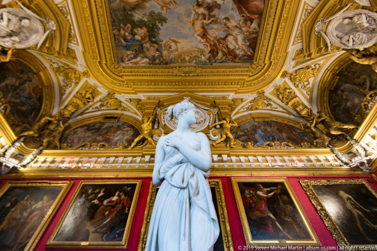 Pale standing female statue seen from behind beneath ornate fresco ceiling and above red walls hung with paintings.