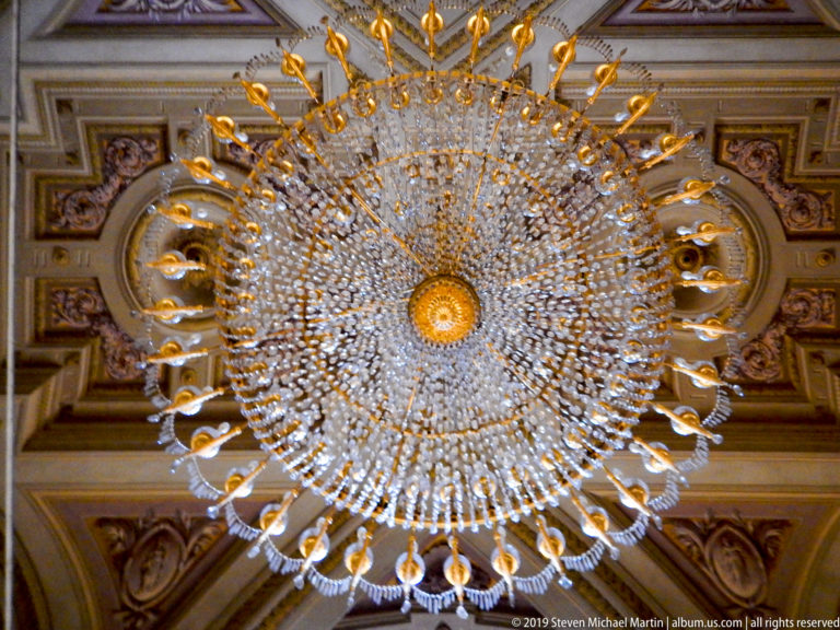 Circular crystal chandelier radiates glass arms and pendants from a golden central hub against a patterned ceiling.