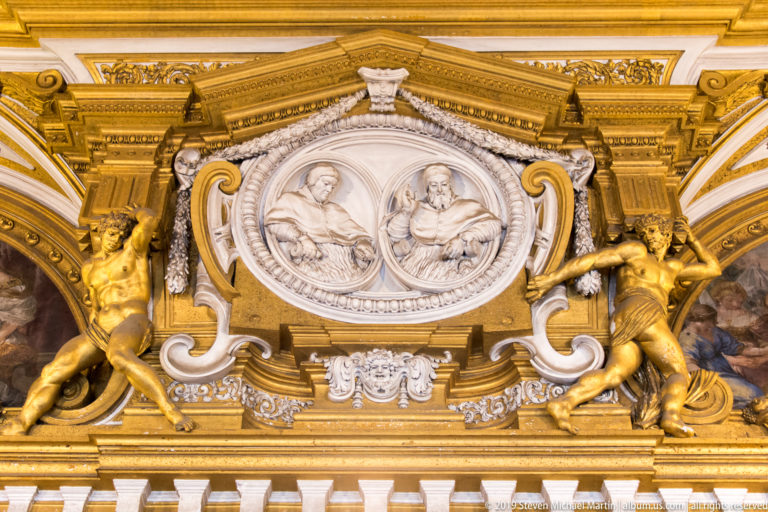 Two seated figures in white oval relief are framed by gold scrollwork and flanked by gilded supporting figures.