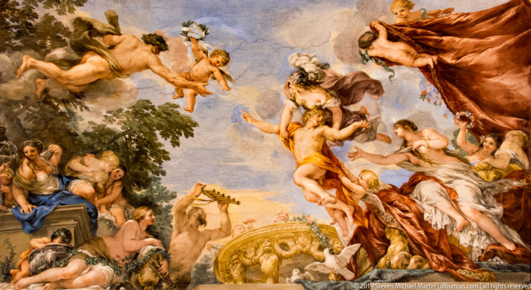 Mythological figures and putti gather in clouds around a central standing figure, with draped fabric and landscape at left.