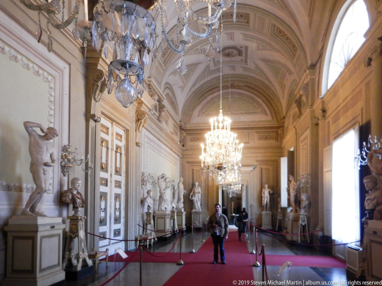 Long decorated palace gallery with sculpture displays, chandeliers, and architectural ornament.