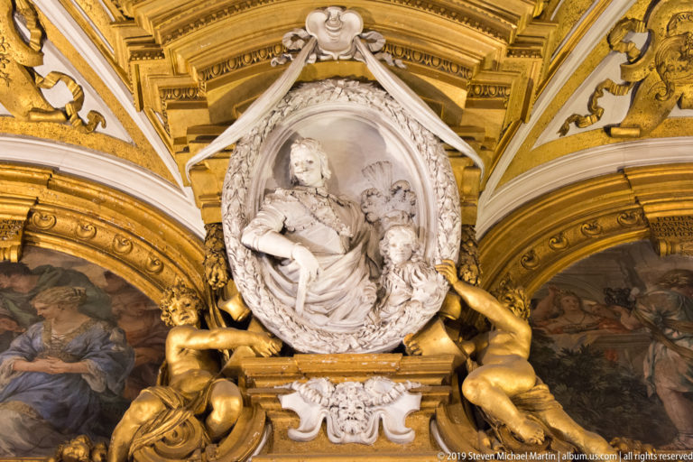 White relief portrait in oval surround is supported by gilded figures amid ceiling paintings and elaborate cornices.
