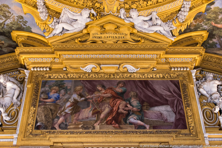 Rectangular painted narrative scene sits below gilded Latin inscription and reclining white stucco figures.
