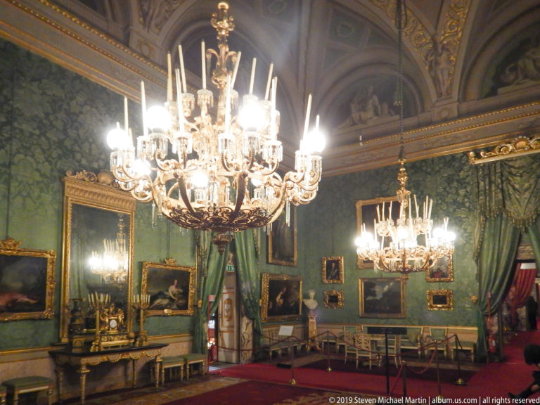 Two lit chandeliers illuminate a long green-walled gallery with gilded framed paintings, mirrors and chairs.