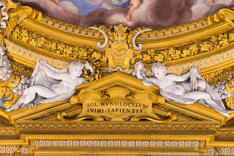 Gold architectural panel with Latin words SOL MUNDI OCULUS ANIMI SAPIENTIA between two reclining pale sculpted figures.
