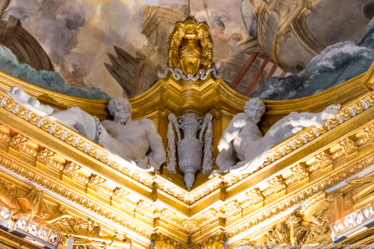 Two pale seated stucco figures flank a central hanging ornament below a gilded lion-like mask and painted clouds.