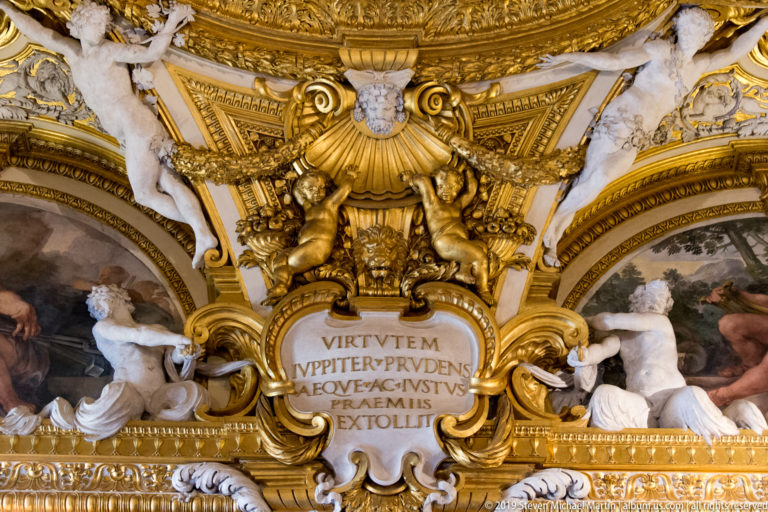 Gold cartouche bears a Latin inscription beneath a sculpted mask, flanked by pale seated figures and ceiling paintings.