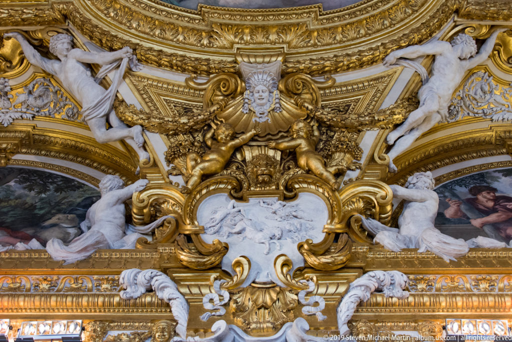 White reclining stucco figures and gilded scrollwork frame an elaborate cartouche below painted ceiling scenes.