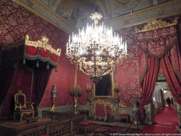 A large chandelier hangs before crimson wall coverings, with a canopy throne at left and curtained doorway at right.
