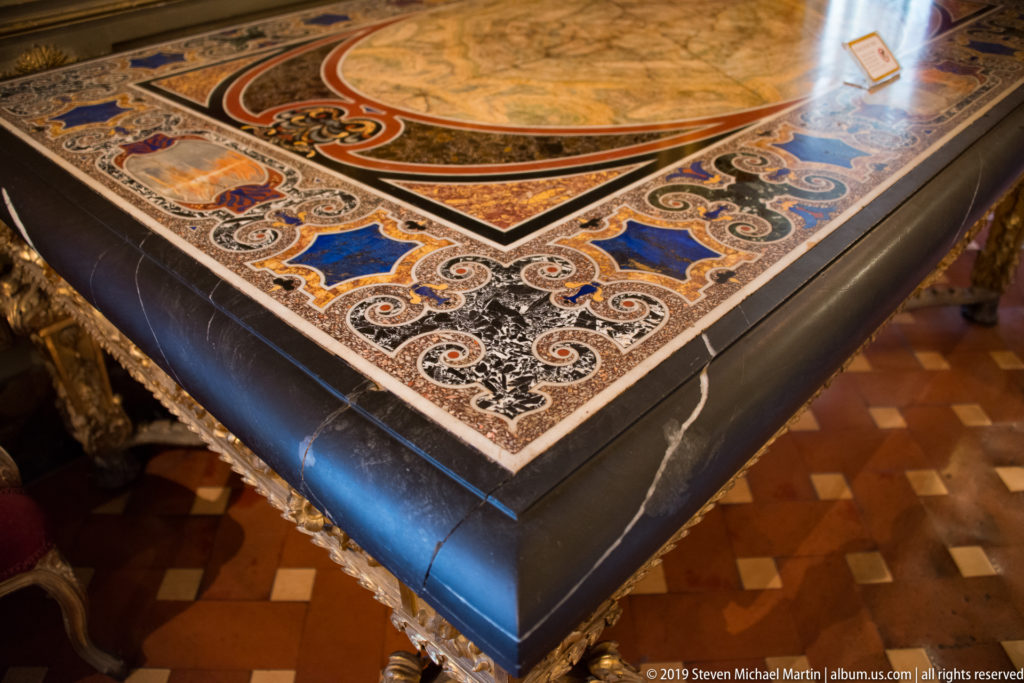 Close view of a dark stone tabletop with colorful scrolling floral inlay, geometric borders and a tiled floor below.
