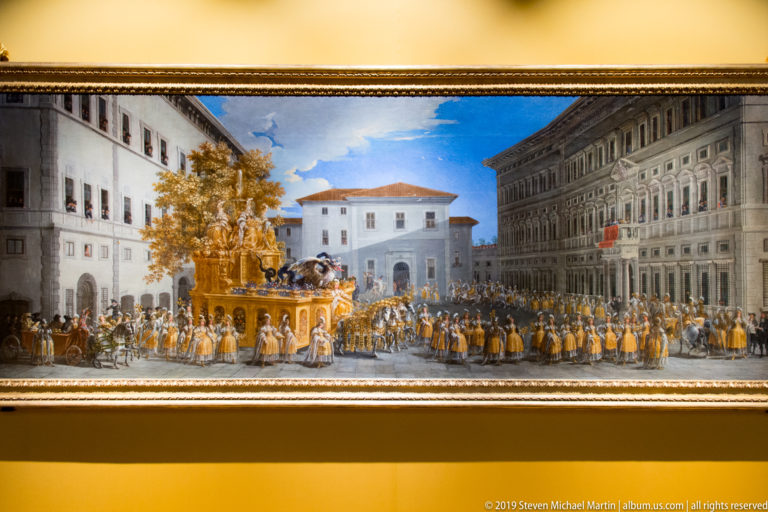 A broad framed painting shows a ceremonial procession with a golden carriage or fountain, ranks of people and a city square.