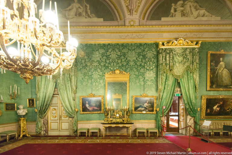 Green damask walls and draperies surround framed paintings, a mantel mirror, chairs and a bright chandelier.