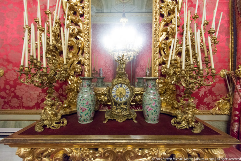 Gilded clock and paired tall candle branches stand before a carved mirror, with painted porcelain vases on a dark console.
