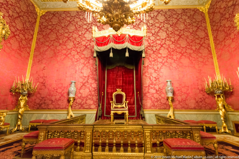 A single gilded red-upholstered chair sits beneath a red canopy on a stepped platform against scarlet patterned walls.