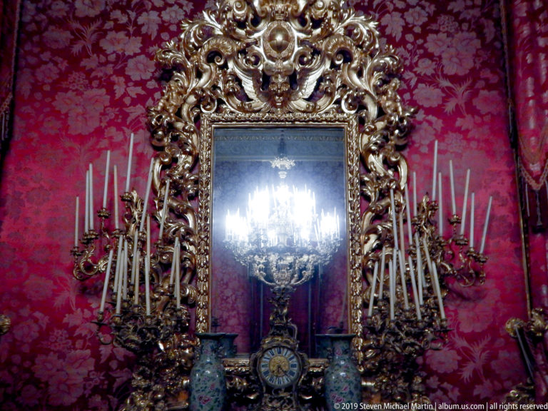 Elaborate gilded mirror above a dark console holds a clock and two floral vases, surrounded by tall candle branches.
