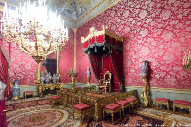 A canopy-covered throne stands on a raised platform behind a gilded railing, with red patterned walls and crystal chandelier.