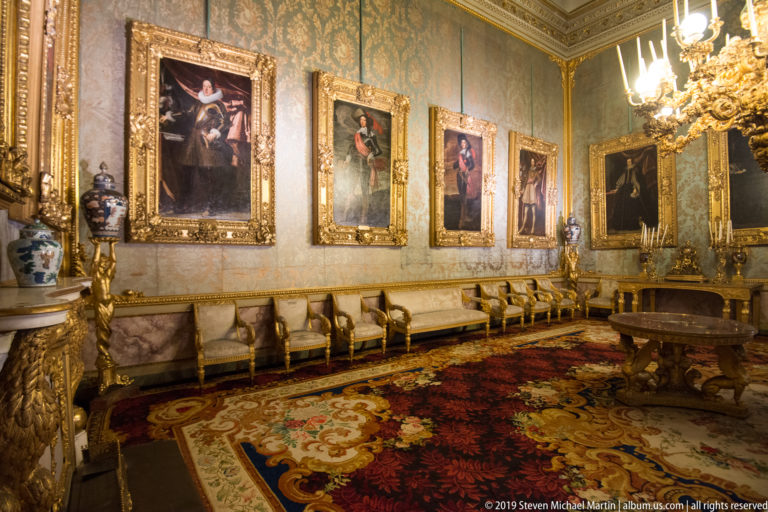 A long reception room has pale patterned walls, gilded portrait frames, upholstered chairs, a chandelier and a floral carpet.