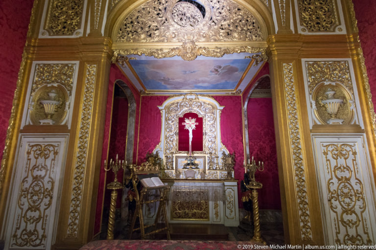 Gilded arched opening frames a small altar beneath a painted blue ceiling, with crimson walls, candlesticks and a radiant emblem.