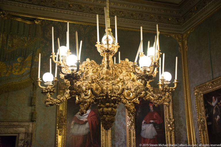 Large ornate gilded chandelier with many lamp arms reflected in a tall mirror inside a richly decorated historic room.