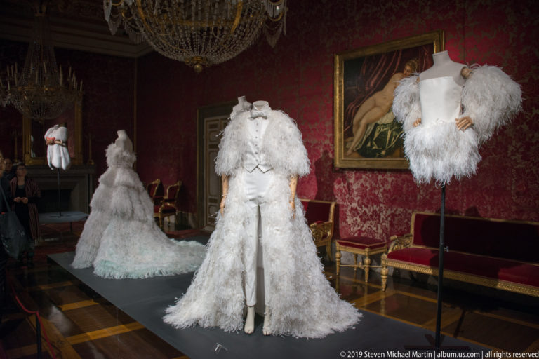 Three white sculptural fashion garments with layered textures and feathers displayed in an ornate red historic gallery.