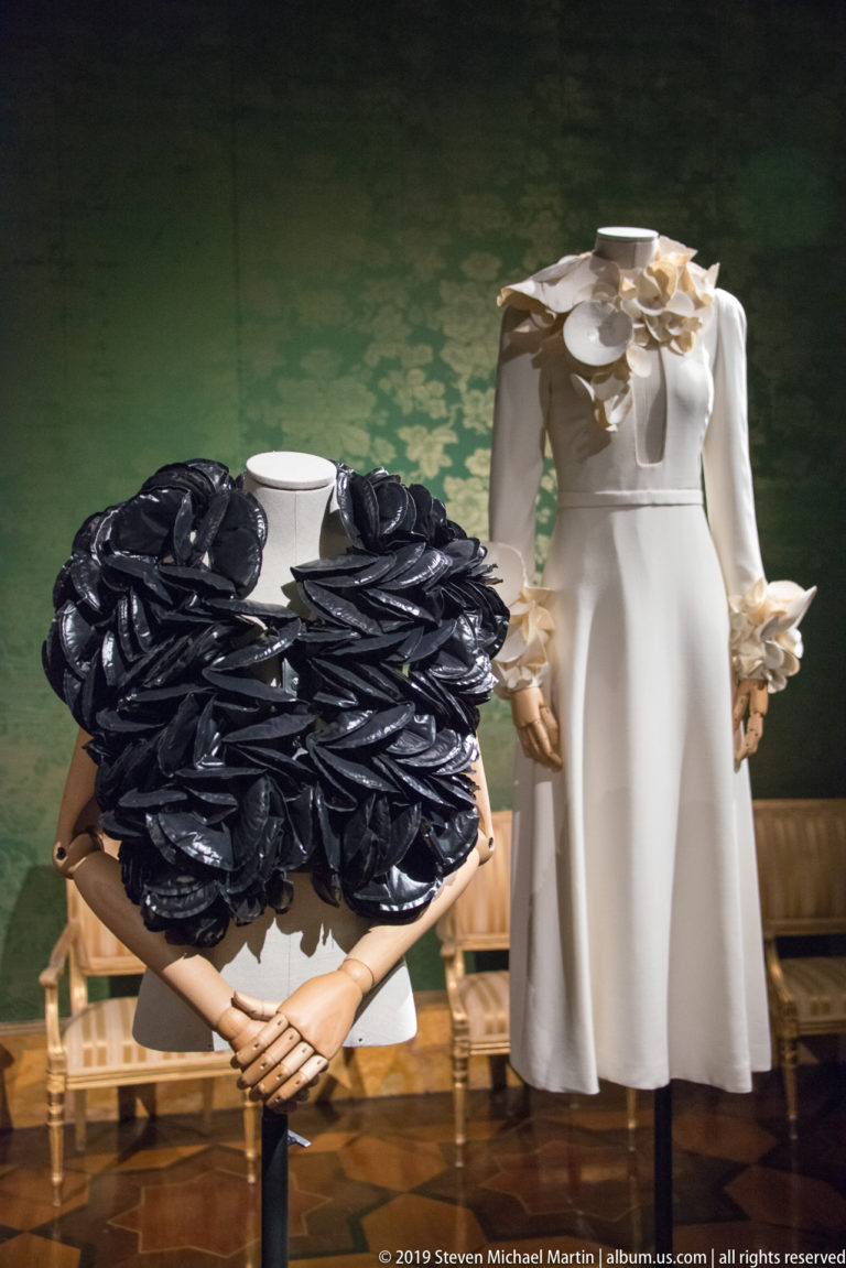 Black garment covered with oversized rosettes beside a white dress with large pale floral forms at the neckline and cuffs.