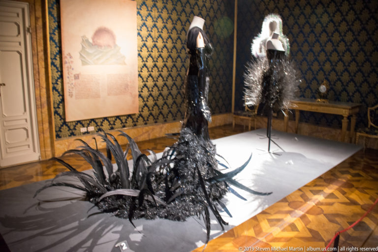 Two black sculptural fashion ensembles with feathered and spiky forms displayed on mannequins in a patterned historic room.