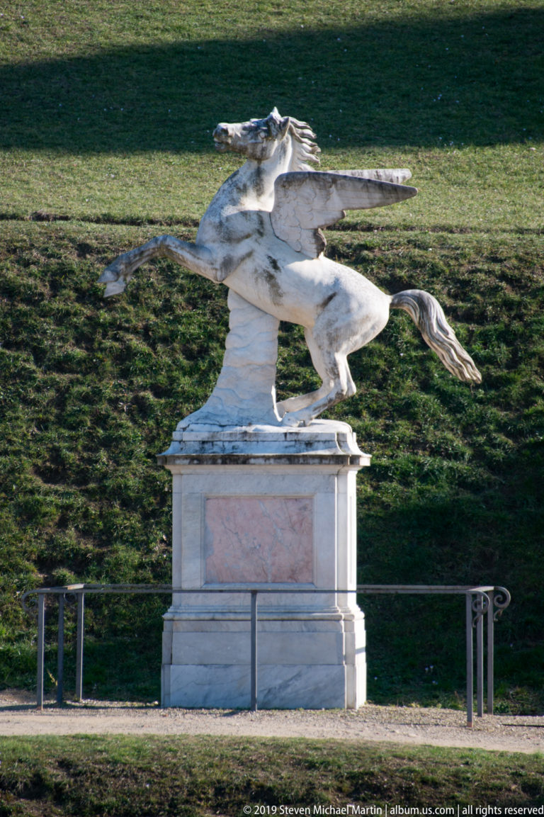 Side view of a white rearing winged horse statue on a pedestal against an open grassy slope.