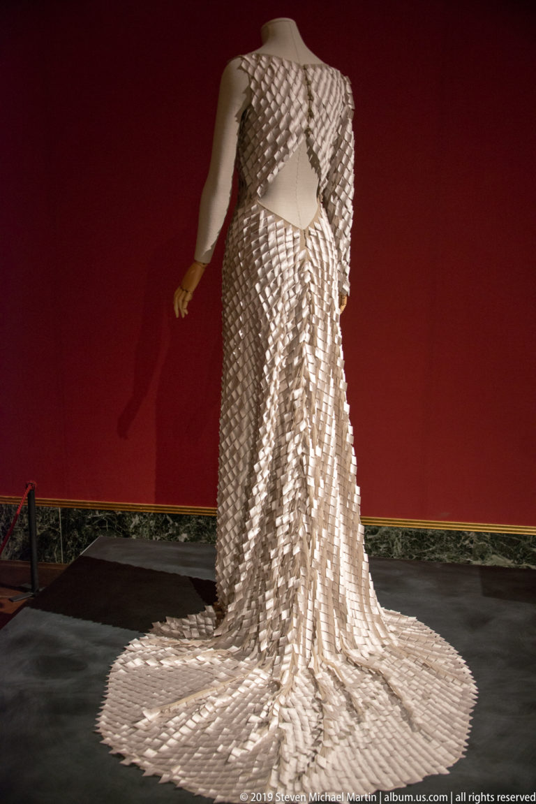 Headless mannequin wearing a reflective silver scale-textured gown with a long circular train against a deep red wall.