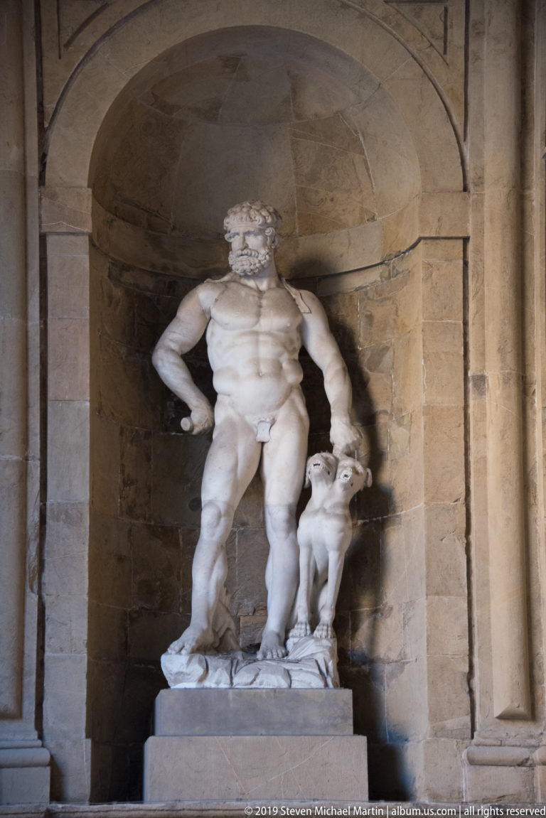 White marble group of a standing muscular nude male with a smaller figure beside him in a stone niche.