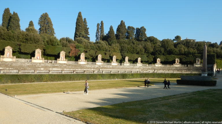 Sunlit lawn bordered by a curved statue-lined stone terrace, trees, visitors, and an Egyptian obelisk at right.