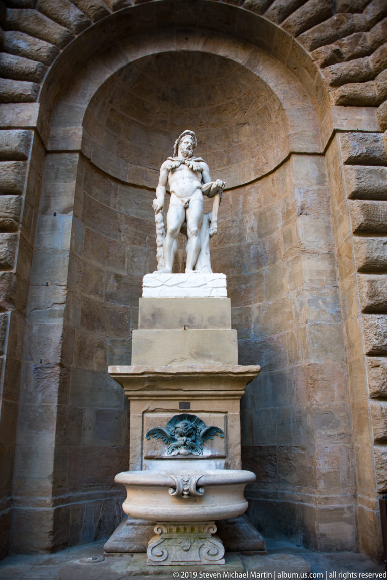 Muscular white marble male statue above a projecting basin inside a deep arched rusticated stone niche.