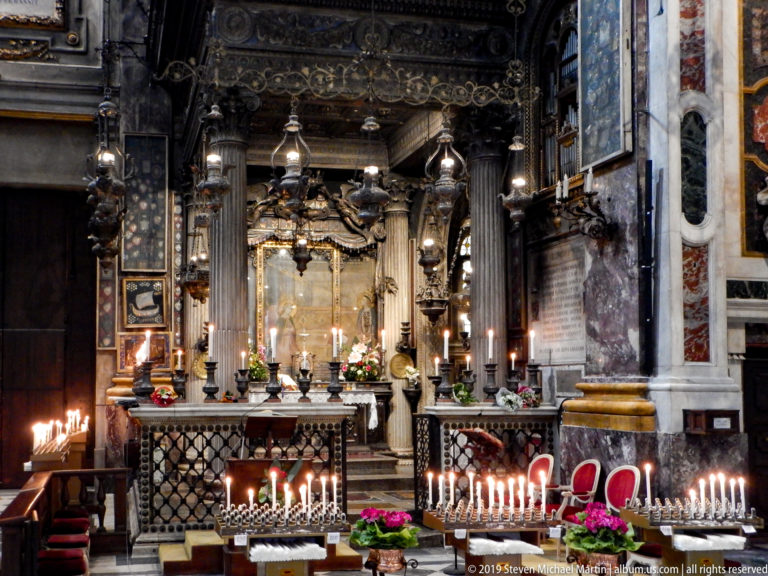 Ornate church altar with votive candles, hanging lamps, flowers, marble piers, and red chairs.