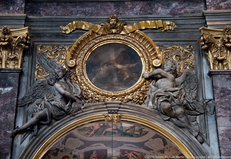Gilded oval painting above an arch, flanked by sculpted figures and elaborate Baroque ornament.
