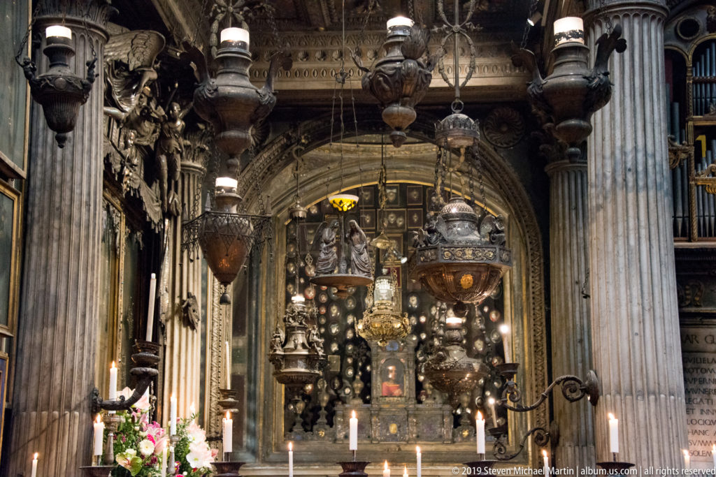 Elaborate devotional shrine surrounded by hanging lamps, candles, flowers, and massive stone columns.