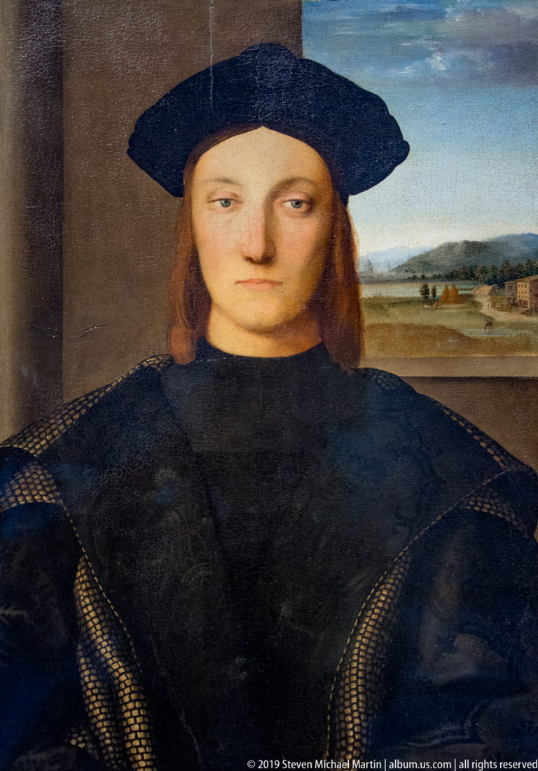 Young man with shoulder-length reddish hair wears a broad black cap and dark clothing before a distant landscape.