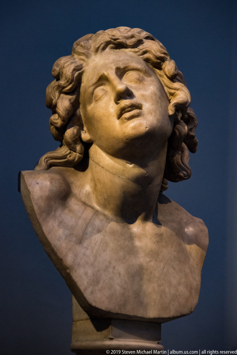 Marble bust of a youthful figure with curling hair, head tilted back and mouth slightly open.