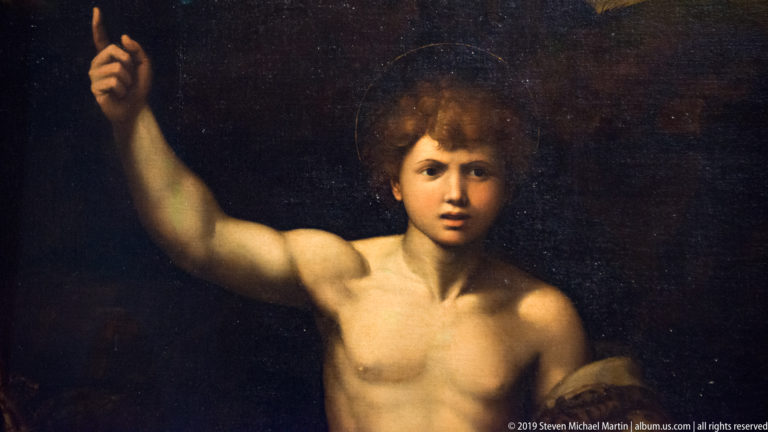 Young nude male figure against a dark background, raising one arm while looking toward the viewer.