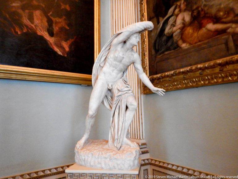 Marble nude male bending forward in a dynamic pose between large framed paintings in the gallery.