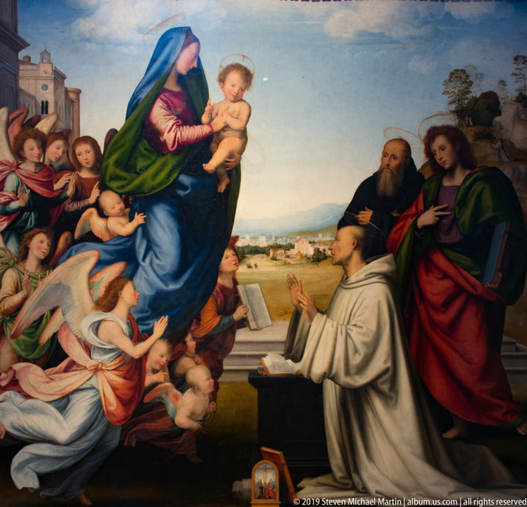 Virgin and Child surrounded by saints and attendants, with a kneeling cleric and open book in the foreground.