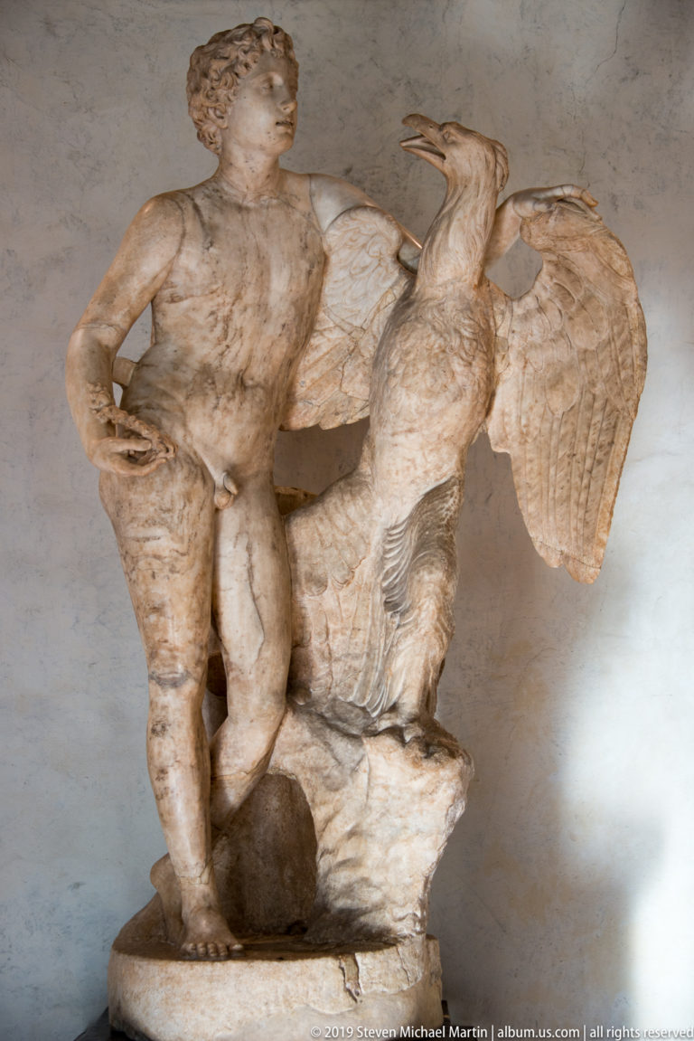 Marble nude male standing beside a large eagle with raised wings in a museum display.