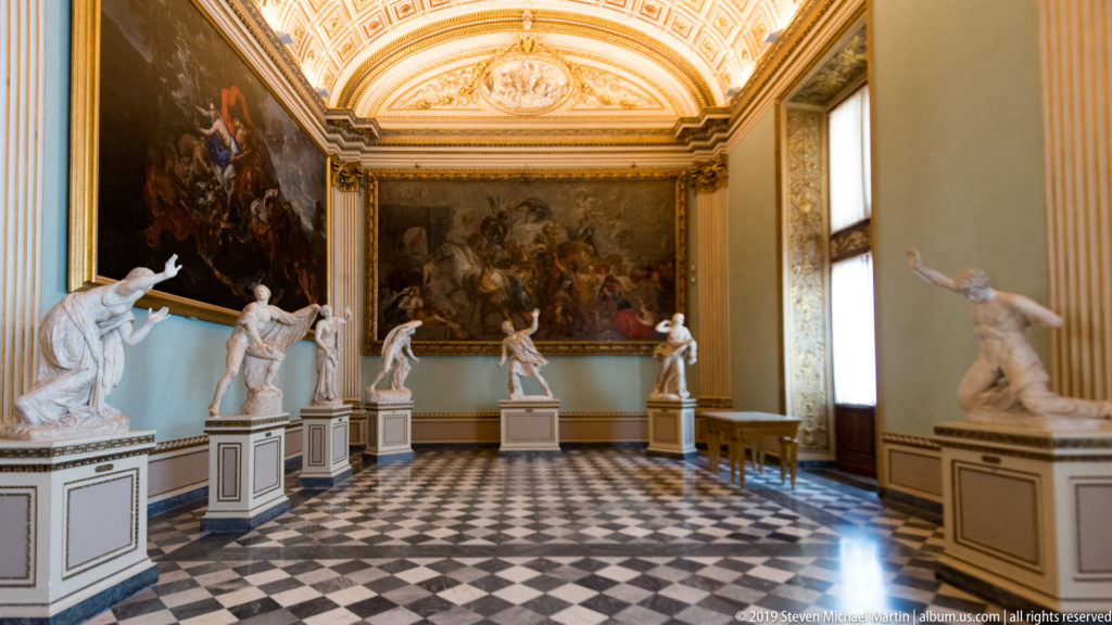 Long Uffizi gallery with marble statues on pedestals, large paintings, checkered floor, and ornate vaulted ceiling.