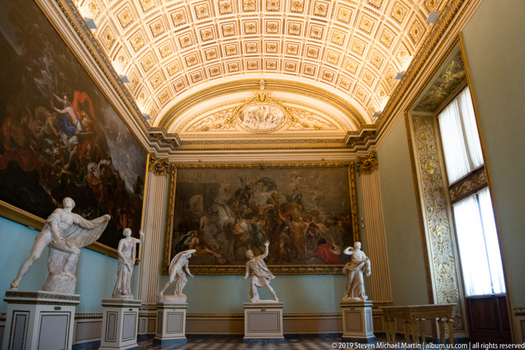 Ornate museum room with marble statues on pedestals, large paintings, tall windows, and a coffered vaulted ceiling.