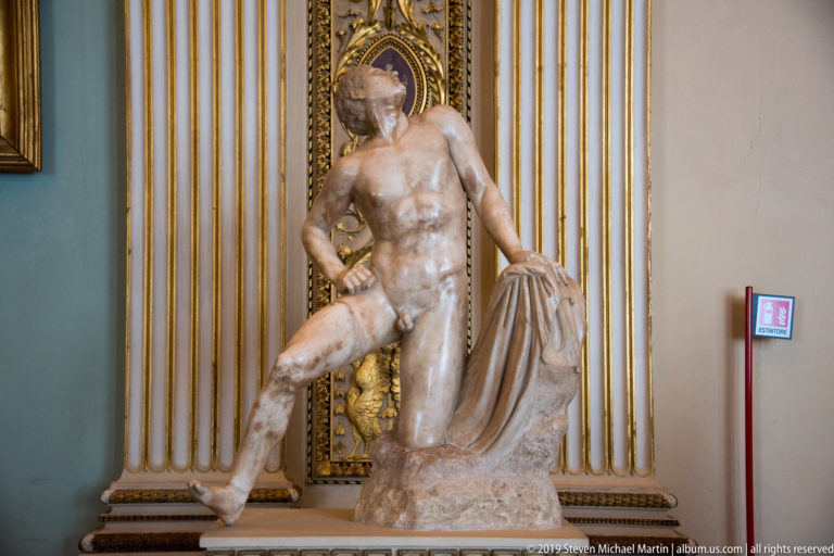 Standing marble nude male with one bent knee, leaning beside a draped support against gilded wall ornament.