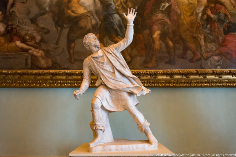 Marble male figure in a short draped garment stepping forward with one hand raised high.