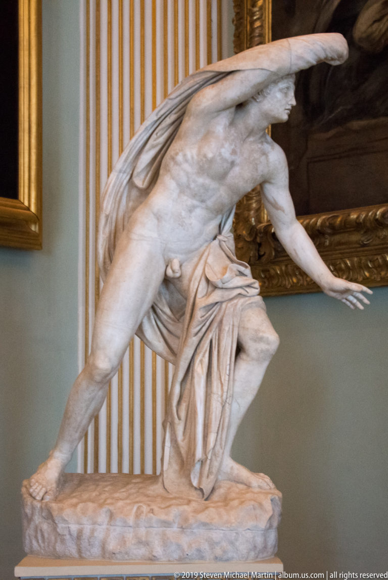 Marble nude male figure bending forward, one arm over his head and drapery gathered behind his legs.