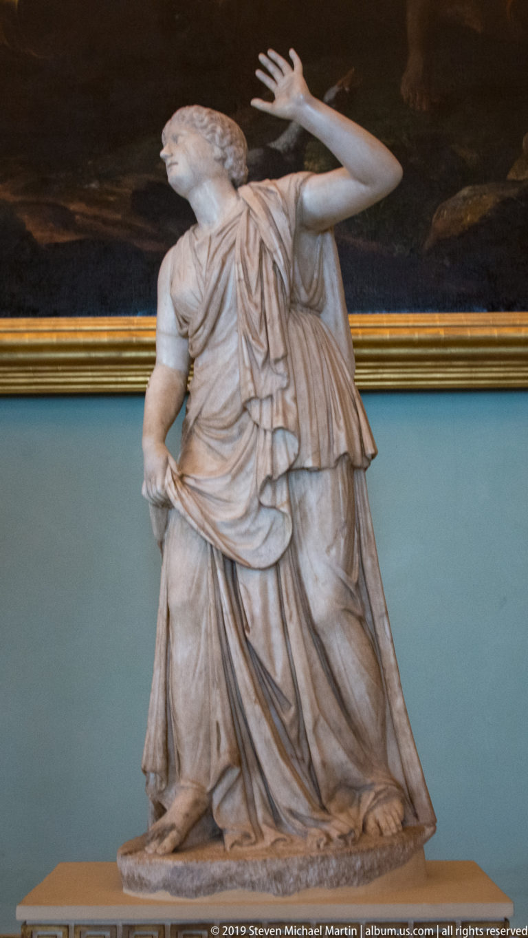 White marble female figure in layered drapery, turning upward with one arm raised beside her head.
