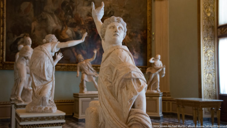 Draped marble female figure raising one arm, with other sculptures and a large painting behind her.